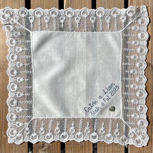 May include: A white handkerchief with a detailed lace border and a small button. The handkerchief has embroidered text that reads "Catrin a Liam 3lain o Fal 2025". The lace trim features a repeating pattern of circles and teardrop shapes.