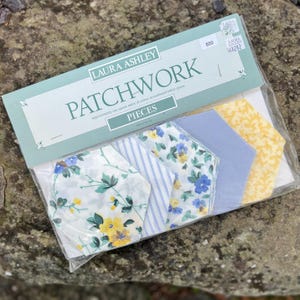 May include: A pack of Laura Ashley patchwork fabric pieces. The pack contains six pieces of fabric in various floral and striped patterns. The fabric is in shades of blue, yellow, and white.