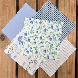 Op de afbeelding: Een assortiment stoffen stalen in verschillende patronen en kleuren. De stoffen omvatten effen lichtblauw, blauw en wit pied-de-poule, een bloemenprint met blauwe bessen en gestreepte en bedrukte stoffen met blauwe en witte accenten. De stalen zijn gerangschikt op een houten oppervlak.