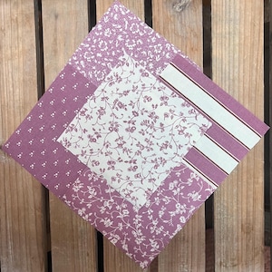 May include: A patchwork quilt square featuring floral and striped patterns in shades of purple and white. The square is made up of several fabric pieces, each with a different design, arranged in a geometric layout. The quilt square is set against a wooden background.