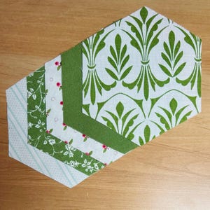 Op de afbeelding: Een verzameling zeshoekige stofstukken in verschillende patronen en kleuren. De stoffen omvatten groen-witte bloemmotieven, een groene streep en effen groen. De stukken zijn gerangschikt op een houten oppervlak.
