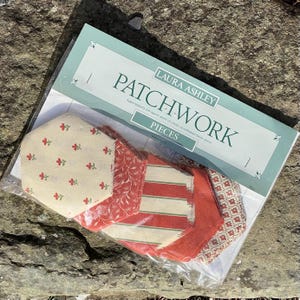 Op de afbeelding: Een pakket met Laura Ashley patchwork-stukken. Het pakket bevat zes zeshoekige stofstukken in rode, witte en groene patronen. Het pakket is gelabeld met "Patchwork Pieces" en "Laura Ashley".