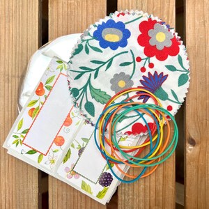 Puede incluir: Una colección de suministros para manualidades que incluye círculos de tela floral, bandas elásticas de colores y etiquetas decorativas con diseños de frutas y flores. La tela presenta flores rojas, azules y grises sobre un fondo blanco. Las etiquetas tienen ilustraciones de naranjas y bayas.