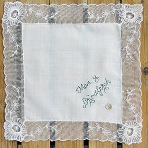 May include: Square white handkerchief with a scalloped lace border adorned with floral patterns. The centre features embroidered text and a small button. The item is displayed on a wooden surface.