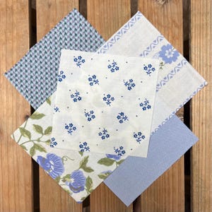 May include: A collection of fabric squares with diverse patterns and colours. The fabrics feature a houndstooth print, floral designs, and solid blue. The squares are arranged on a wooden surface, displaying the different textures and designs.