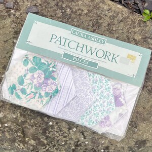 Pode incluir: Um pacote de pe&ccedil;as de patchwork Laura Ashley. O pacote cont&eacute;m v&aacute;rios peda&ccedil;os de tecido com desenhos florais e estampados em tons de roxo, verde e branco. O pacote &eacute; selado em pl&aacute;stico transparente.