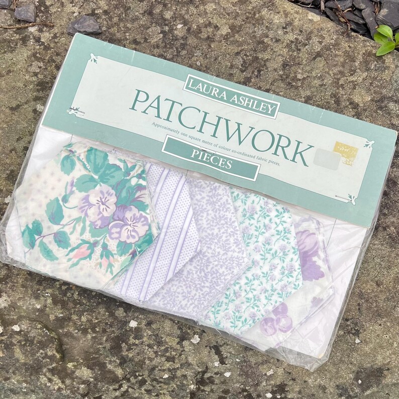 Pode incluir: Um pacote de pe&ccedil;as de patchwork Laura Ashley. O pacote cont&eacute;m v&aacute;rios peda&ccedil;os de tecido com desenhos florais e estampados em tons de roxo, verde e branco. O pacote &eacute; selado em pl&aacute;stico transparente.