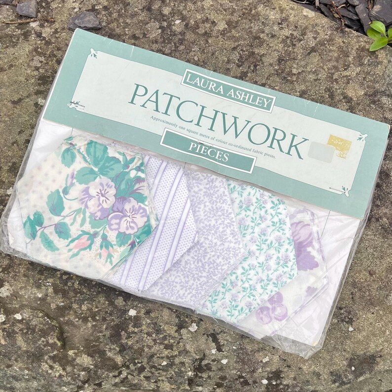 Pode incluir: Um kit de patchwork Laura Ashley com v&aacute;rias pe&ccedil;as de tecido com motivos florais e estampados. O pacote inclui tecidos em tons de branco, roxo e verde-azulado. A embalagem diz "PATCHWORK" e "PIECES".