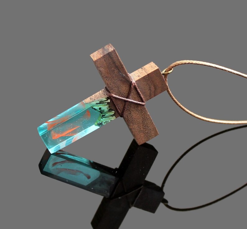 May include: A wooden cross pendant with a blue and orange resin inlay. The pendant is attached to a brown cord.