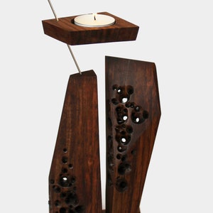 Peut inclure: Un bougeoir en bois avec un design g&eacute;om&eacute;trique. Le bougeoir a un petit porte-bougie rond sur le dessus et deux pi&egrave;ces rectangulaires hautes sur le dessous. Le bois est brun fonc&eacute; et a une finition naturelle et rustique.