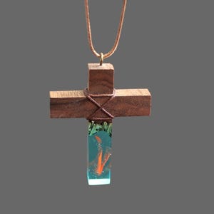 May include: A wooden cross necklace with a teal blue resin pendant containing a small orange swirl. The cross is wrapped with brown twine.