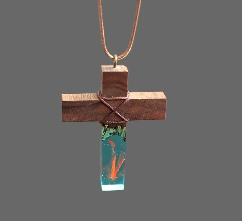 May include: A wooden cross necklace with a teal blue resin pendant containing a small orange swirl. The cross is wrapped with brown twine.
