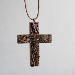 May include: A rustic wooden cross pendant with a textured, bark-like appearance. The cross is suspended from a brown cord, creating a simple yet striking piece of jewelry. The pendant's natural wood grain adds to its unique charm.