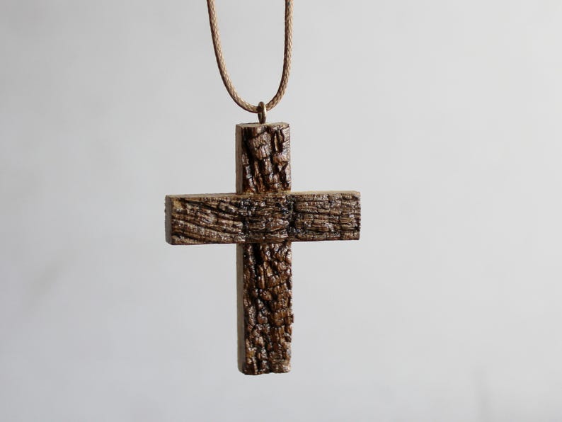 May include: A rustic wooden cross pendant with a textured, bark-like appearance. The cross is suspended from a brown cord, creating a simple yet striking piece of jewelry. The pendant's natural wood grain adds to its unique charm.
