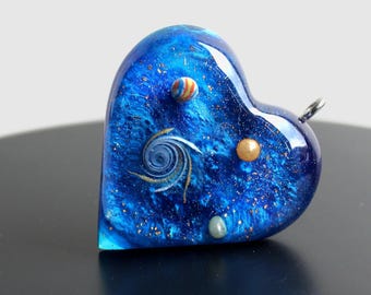 May include: A blue heart-shaped pendant with a galaxy design, featuring a swirling nebula, gold glitter, and small colorful planets.