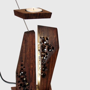 Peut inclure: Un bougeoir en bois avec un design g&eacute;om&eacute;trique. Le bougeoir a un petit porte-bougie rond sur le dessus et une base rectangulaire plus grande avec un motif sculpt&eacute;. Le bougeoir est allum&eacute; et la lumi&egrave;re brille &agrave; travers le motif sculpt&eacute;.