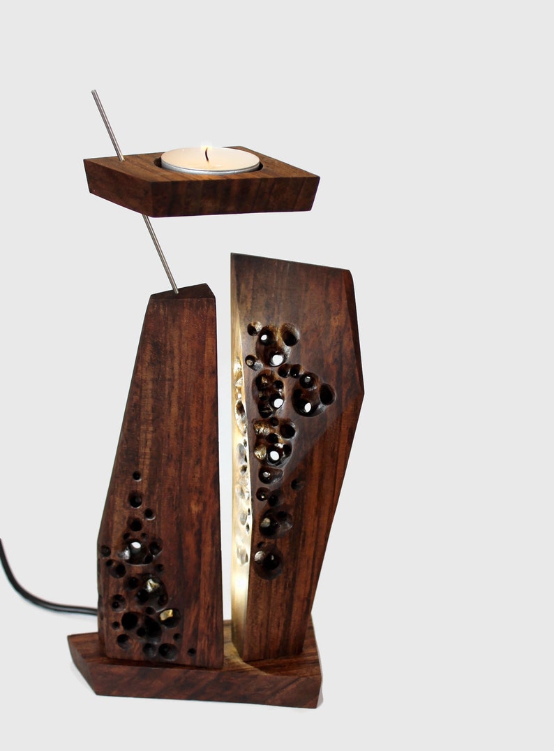Peut inclure: Un bougeoir en bois avec un design g&eacute;om&eacute;trique. Le bougeoir a un petit porte-bougie rond sur le dessus et une base rectangulaire plus grande avec un motif sculpt&eacute;. Le bougeoir est allum&eacute; et la lumi&egrave;re brille &agrave; travers le motif sculpt&eacute;.