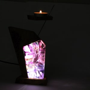Peut inclure: Une lampe en bois unique avec un design g&eacute;om&eacute;trique. La lampe pr&eacute;sente un int&eacute;rieur abstrait et lumineux avec des nuances de violet et de blanc. Un petit bougeoir avec une bougie allum&eacute;e flotte au-dessus de la lampe, reli&eacute; par une fine tige m&eacute;tallique.