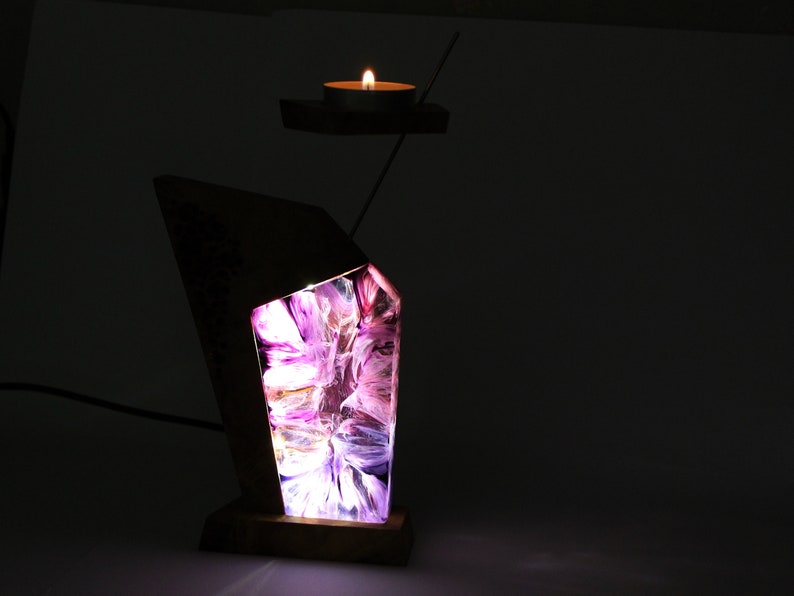 Peut inclure: Une lampe en bois unique avec un design g&eacute;om&eacute;trique. La lampe pr&eacute;sente un int&eacute;rieur abstrait et lumineux avec des nuances de violet et de blanc. Un petit bougeoir avec une bougie allum&eacute;e flotte au-dessus de la lampe, reli&eacute; par une fine tige m&eacute;tallique.