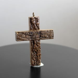 May include: A handcrafted wooden cross with a textured, bark-like appearance. The cross is brown with a small metal loop at the top for hanging. It stands on a dark surface, suggesting a decorative or religious item.