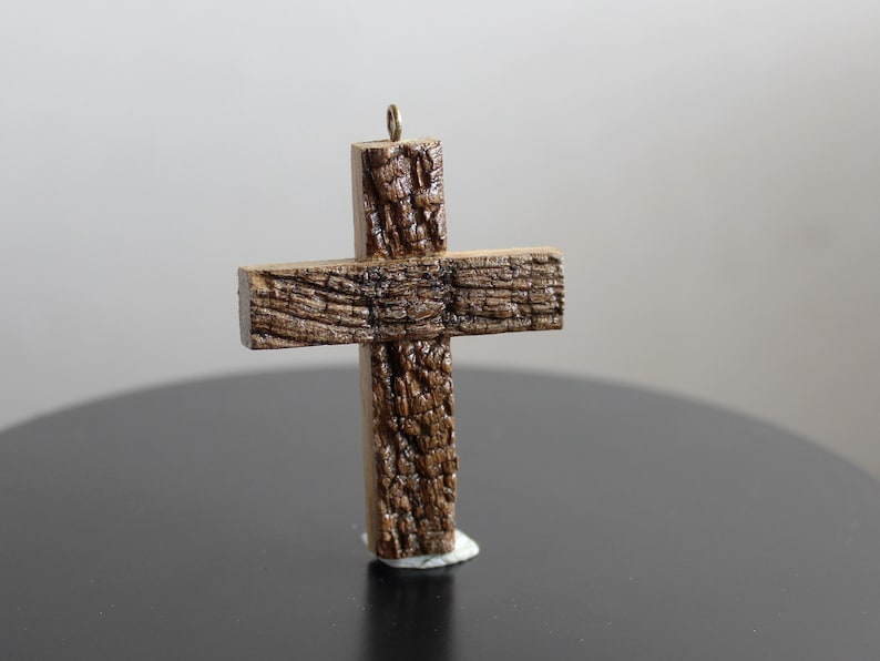 May include: A handcrafted wooden cross with a textured, bark-like appearance. The cross is brown with a small metal loop at the top for hanging. It stands on a dark surface, suggesting a decorative or religious item.