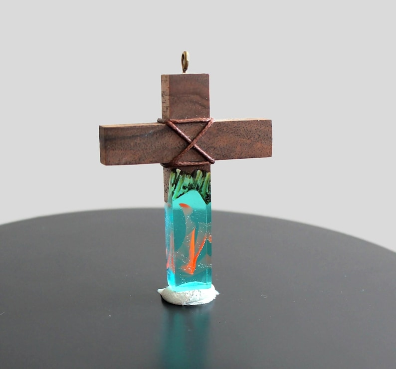 May include: A wooden cross with a blue and green resin center. The resin has orange swirls inside. The cross is tied with brown twine.