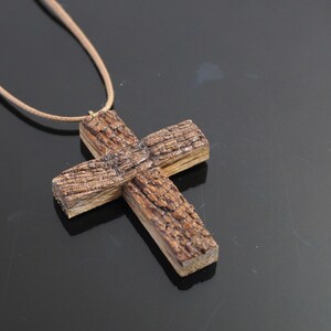 May include: A handcrafted wooden cross pendant on a tan cord. The cross is made of textured, brown wood, with visible grain and a rustic appearance. The necklace is displayed against a dark background.