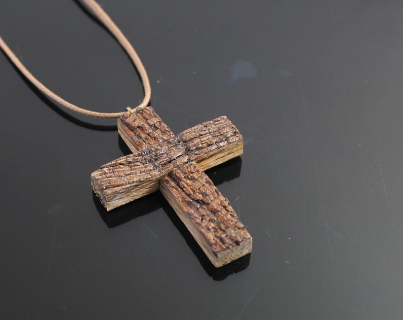 May include: A handcrafted wooden cross pendant on a tan cord. The cross is made of textured, brown wood, with visible grain and a rustic appearance. The necklace is displayed against a dark background.