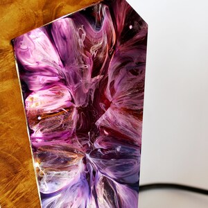 Peut inclure: Gros plan sur un objet d&eacute;coratif en bois avec un design abstrait. Il pr&eacute;sente une section de bois brun clair avec des taches sombres et une section de r&eacute;sine tourbillonnante violette, rose et blanche. Un cordon noir est visible.
