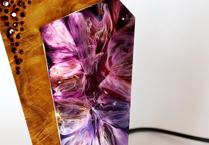 Peut inclure: Gros plan sur un objet d&eacute;coratif en bois avec un design abstrait. Il pr&eacute;sente une section de bois brun clair avec des taches sombres et une section de r&eacute;sine tourbillonnante violette, rose et blanche. Un cordon noir est visible.