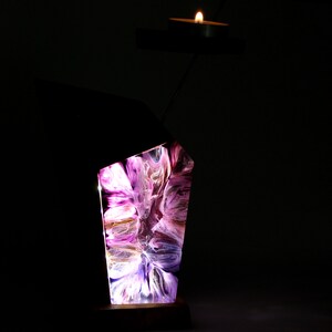 Peut inclure: Un bougeoir d&eacute;coratif avec un design abstrait. Le support pr&eacute;sente un bloc de r&eacute;sine rectangulaire et &eacute;clair&eacute; avec des couleurs tourbillonnantes violettes, blanches et dor&eacute;es. Une petite bougie est pos&eacute;e sur un bras en m&eacute;tal noir, projetant une lueur chaleureuse. La base est en bois fonc&eacute;.