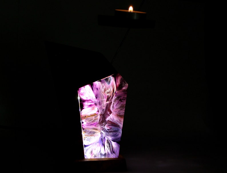 Peut inclure: Un bougeoir d&eacute;coratif avec un design abstrait. Le support pr&eacute;sente un bloc de r&eacute;sine rectangulaire et &eacute;clair&eacute; avec des couleurs tourbillonnantes violettes, blanches et dor&eacute;es. Une petite bougie est pos&eacute;e sur un bras en m&eacute;tal noir, projetant une lueur chaleureuse. La base est en bois fonc&eacute;.