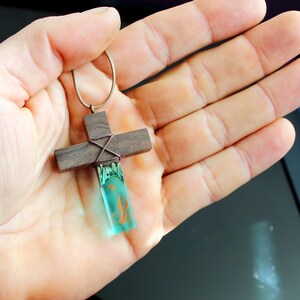 May include: A wooden cross pendant with a teal blue resin inlay. The cross is wrapped with brown cord and has a brown leather cord attached.