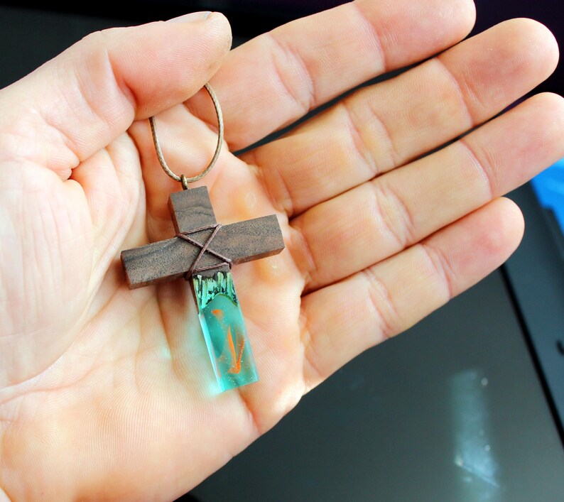 May include: A wooden cross pendant with a teal blue resin inlay. The cross is wrapped with brown cord and has a brown leather cord attached.