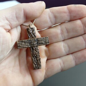 May include: A handcrafted wooden cross pendant with a textured, bark-like appearance. The cross is suspended from a thin, light-brown cord. The pendant is held in an open hand, showcasing its rustic design and natural wood grain.