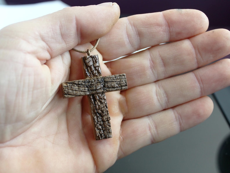 May include: A handcrafted wooden cross pendant with a textured, bark-like appearance. The cross is suspended from a thin, light-brown cord. The pendant is held in an open hand, showcasing its rustic design and natural wood grain.