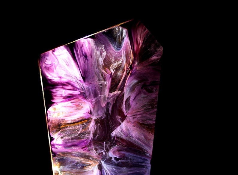 Peut inclure: Pi&egrave;ce d'art abstraite en r&eacute;sine avec des pigments violets, blancs et dor&eacute;s tourbillonnants. L'&oelig;uvre est enferm&eacute;e dans une forme g&eacute;om&eacute;trique claire, cr&eacute;ant un effet visuel unique. Le fond est noir uni.