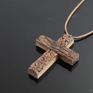 May include: A wooden cross pendant with a textured, weathered appearance. The cross is made of brown wood and is suspended from a tan cord. The cross is approximately 3 inches wide and 4 inches long.