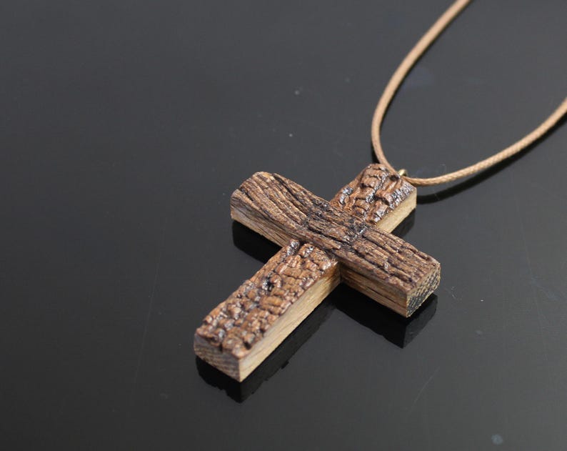 May include: A wooden cross pendant with a textured, weathered appearance. The cross is made of brown wood and is suspended from a tan cord. The cross is approximately 3 inches wide and 4 inches long.