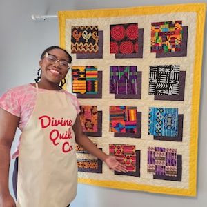 Może przedstawiać: Osoba w białym fartuchu z napisem "Divine Quilt Co." w czerwonych literach, stojąca przed kołdrą z żółtą obwódką i 12 kolorowymi kwadratami z geometrycznymi wzorami.