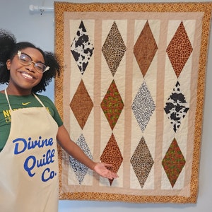 Op de afbeelding: Een persoon draagt een groen shirt en een beige schort met de tekst "Divine Quilt Co." en staat voor een quilt met een ruitpatroon. De quilt is gemaakt van verschillende stoffen met dierenprintontwerpen, waaronder koehuid, luipaard en giraffe.