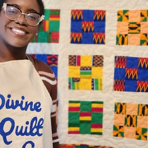 Può includere: Una donna indossa una camicia a righe marroni e bianche e un grembiule bianco con il testo "Divine Quilt Co." in lettere blu. È in piedi davanti a una trapunta bianca con quadrati colorati che presentano tessuti stampati africani.