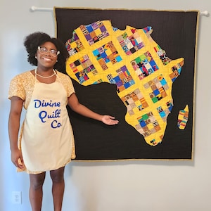 Könnte beinhalten: Eine große, farbenfrohe Steppdecke in Form des afrikanischen Kontinents. Die Steppdecke hat einen gelben Hintergrund mit einem Patchwork-Design aus verschiedenen Stoffmustern. Die Steppdecke wird auf einem schwarzen Hintergrund mit einem goldenen Rand präsentiert. Ein Schürze mit dem Text "Divine Quilt Co." ist ebenfalls sichtbar.