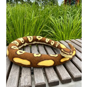 Könnte beinhalten: Eine braune und gelbe Filzschlange, die sich auf einer Holzbank zu einem Kreis zusammenrollt. Die Schlange hat eine realistische Textur und ist ein handgemachtes Handwerk.