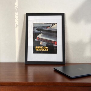 May include: A black framed wall art print featuring a rear view of a silver Porsche sports car with a prominent spoiler and taillights. The text "SAVE the WHALES" is printed in bold yellow letters at the bottom of the image within the frame.