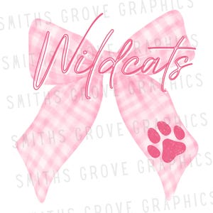 Puede incluir: Un lazo rosa a cuadros con la palabra "Wildcats" en cursiva. Una huella de pata rosa está en el lado inferior derecho del lazo. El lazo es un diseño gráfico, probablemente para ropa o accesorios.