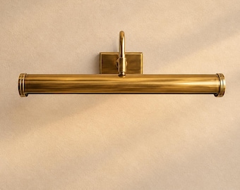 May include: A horizontal wall-mounted picture light fixture with a brushed brass finish. The lamp features a long cylindrical tube attached to a rectangular wall plate by a single curved arm, creating a sleek and functional lighting design.