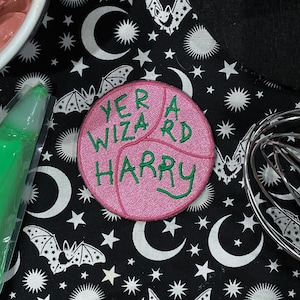 Peut inclure: Un écusson rose avec la phrase "Yer a Wizard Harry" brodée en vert. L'écusson est posé sur un tissu noir orné de motifs blancs de lunes, d'étoiles et de chauves-souris. Un bol de glaçage rose, une poche à douille verte et un chapeau noir sont également visibles.