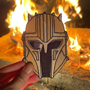 May include: A gold and black helmet with a detailed design, held in front of a fire pit.