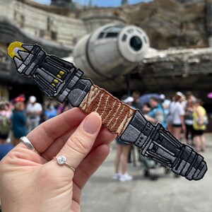 May include: A detailed patch of a lightsaber, featuring a gray and black hilt with yellow accents and a brown grip. The patch is held up in front of a blurred background of a Star Wars-themed park, with the Millennium Falcon visible.